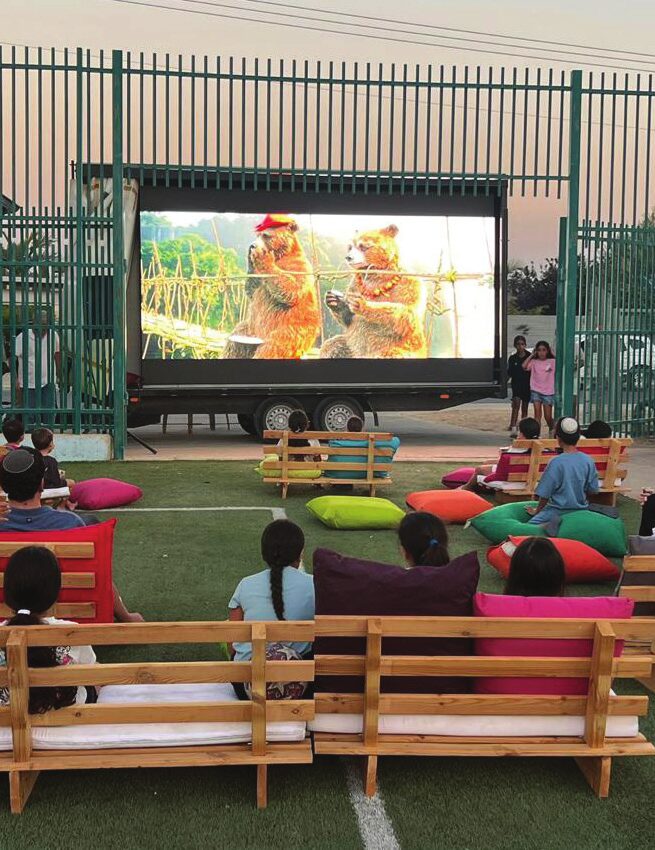 הקולנוע הנייד — מסך גדול באוויר הפתוח וקהל יושב על ספסלים וכריות